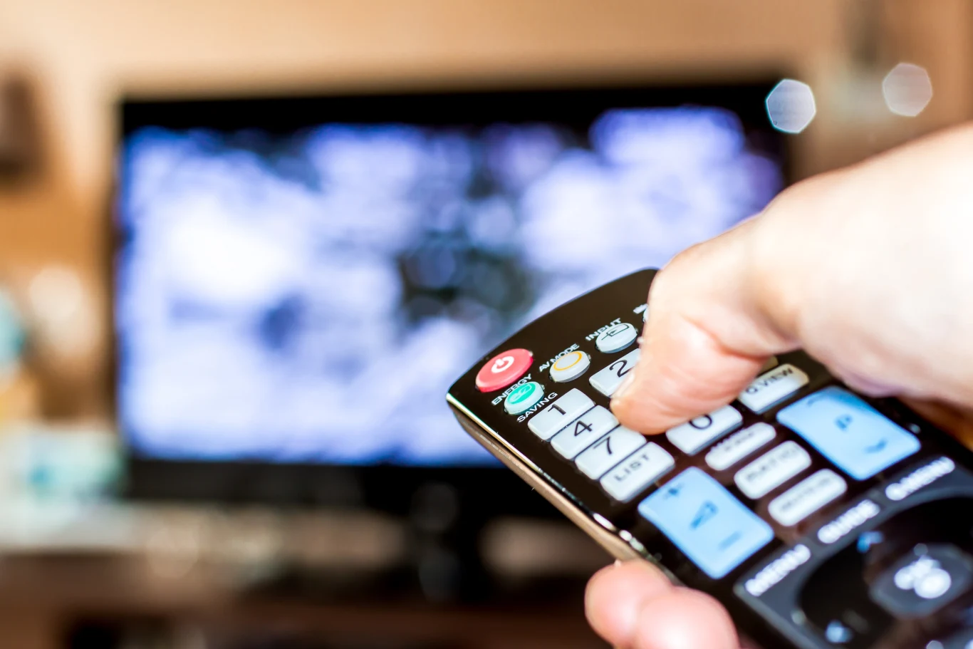 Nowa opłata audiowizualna zamiast abonamentu RTV Pilot trzymany w dłoni skierowany w stronę włączonego telewizora; nowe zasady opłaty audiowizualnej