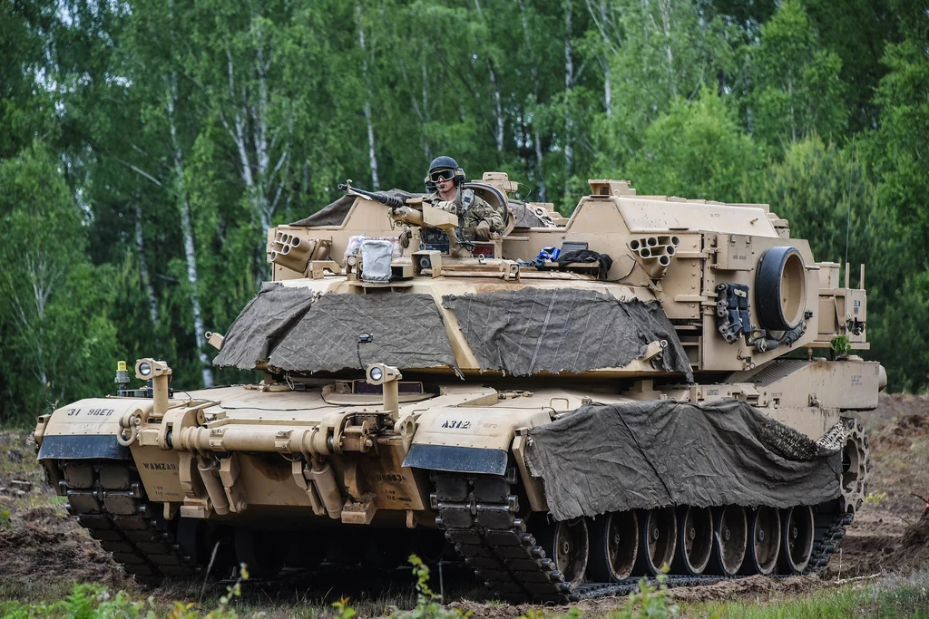 Amerykański M1150 na poligonie w Drawsku Pomorskim w 2020 roku Czołg wojskowy poruszający się po terenie leśnym, wyposażony w dodatkowe osłony i akcesoria, z widocznym żołnierzem siedzącym w wieżyczce, otoczony zielenią lasu.