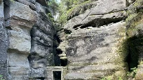 Wąska ścieżka prowadząca przez drewnianą kładkę i kamienną bramę między wysokimi, naturalnymi skałami, otoczona gęstą roślinnością, w tym paprociami, tworząc atmosferę tajemniczości i dzikości.