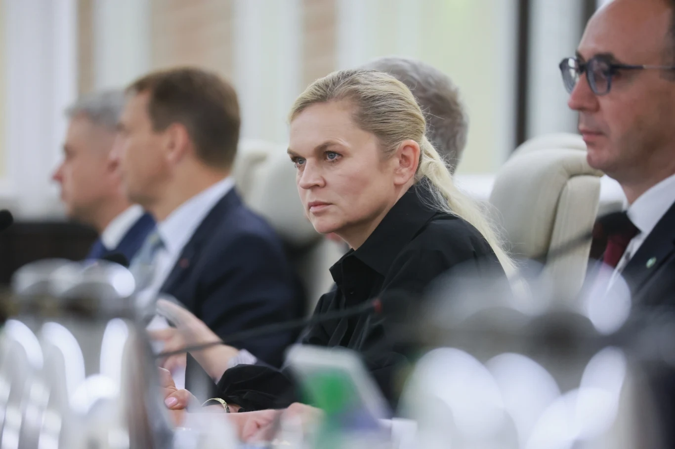 Minister edukacji Barbara Nowacka Kobieta o blond włosach siedząca przy stole konferencyjnym, skupiona na rozmowie, otoczona przez innych uczestników spotkania w formalnych garniturach.