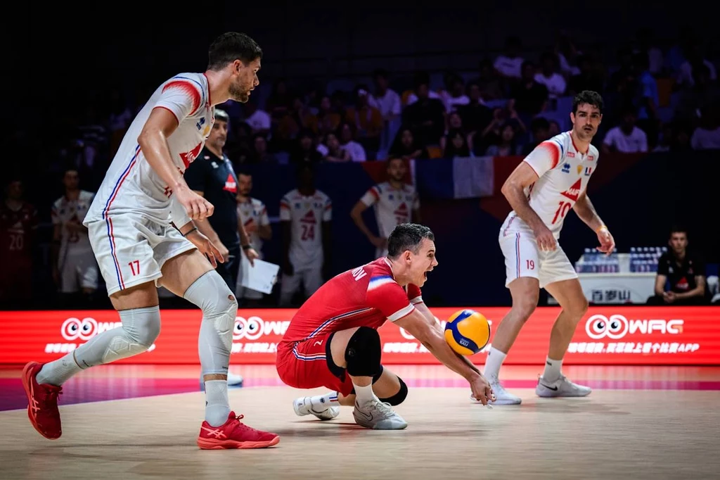 Liga Narodów siatkarzy. Francja miała problemy ze Słowenią Zawodnik drużyny siatkarskiej wykonuje odbiór piłki podczas intensywnej akcji na boisku, otoczony przez dwóch współgraczy w białych strojach, w tle widoczna publiczność oraz elementy hali sportowej.