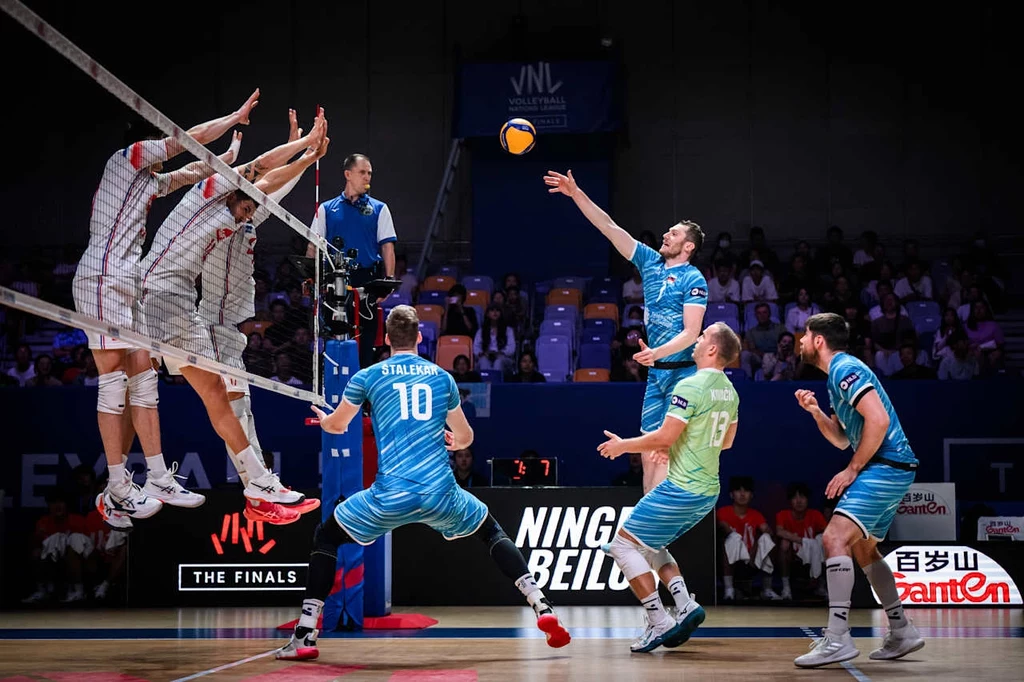 Liga Narodów siatkarzy. Kadr z meczu Francja - Słowenia Mecz siatkówki halowej z wyraźnym momentem ataku drużyny w niebieskich strojach, trzech zawodników drużyny przeciwnej skacze w bloku przy siatce, sędzia obserwuje grę z podwyższenia, kibice w tle.