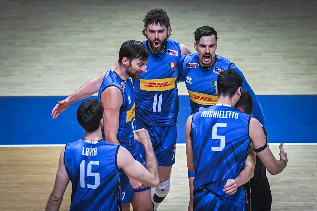 Grupa siatkarzy w niebieskich strojach z włoskimi flagami na ramieniu świętuje zdobycie punktu, widoczne emocje oraz zgranie drużynowe, na parkiecie hali sportowej.