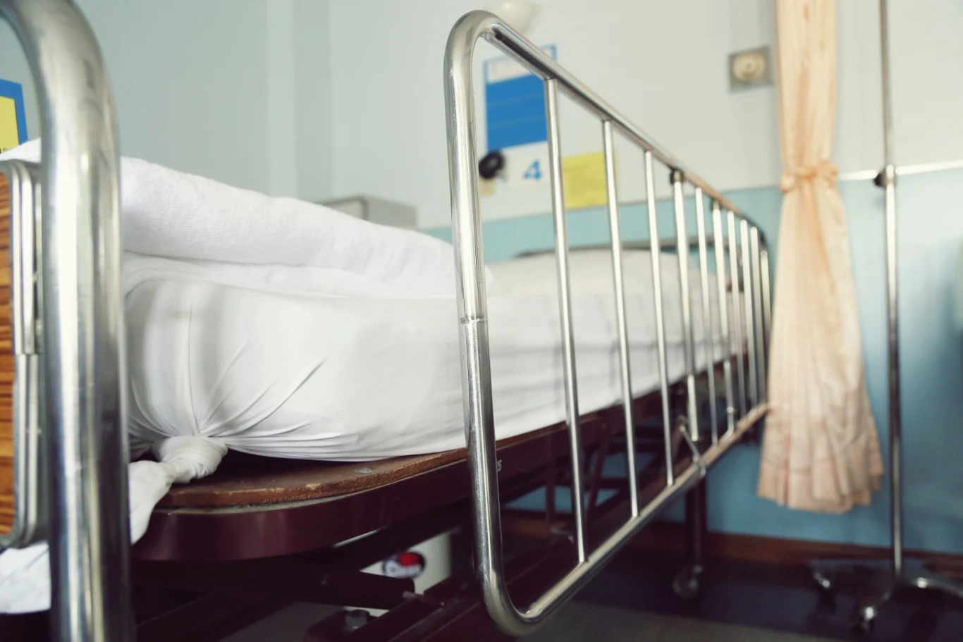 Choroba legionistów w USA. Zmarła jedna osoba, zakażenie u kilkudziesięciu innych Łóżko szpitalne z metalowymi barierkami, świeżo pościelone, w tle pastelowe ściany, żółta zasłona oraz stojak medyczny.