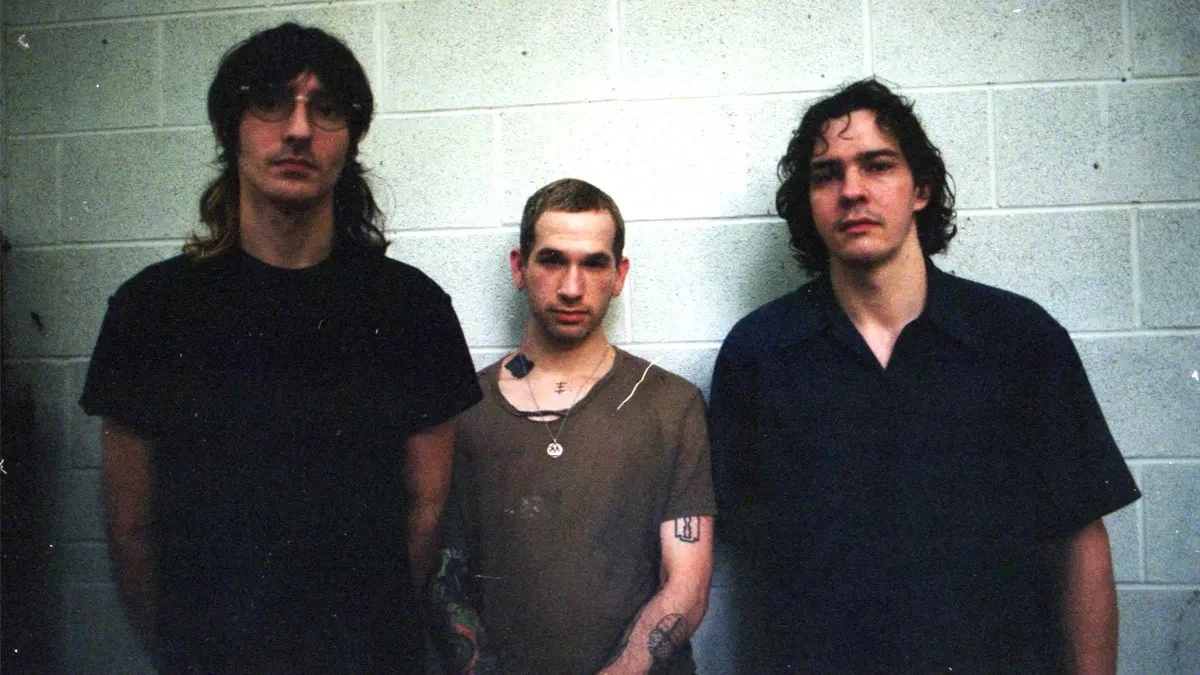 Portrayal of Guilt Trzech mężczyzn stoi na tle jasnej ściany z cegieł, środkowy mężczyzna ma widoczne tatuaże na rękach i szyi, wszyscy patrzą prosto w obiektyw, ubrani są w ciemne, casualowe ubrania.