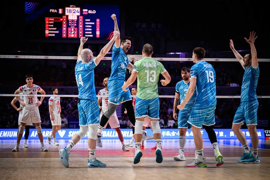 Mecz Francja - Słowenia w Lidze Narodów Grupa siatkarzy w niebieskich i zielonych strojach świętuje zdobycie punktu podczas meczu przeciwko drużynie w białych strojach, w tle widoczna tablica wyników oraz kibice.