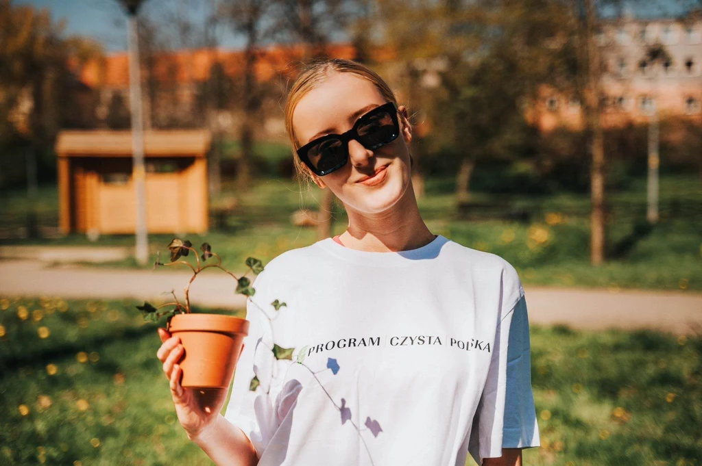 Uśmiechnięta młoda kobieta w ciemnych okularach przeciwsłonecznych, trzymająca w ręku doniczkę z rośliną; nosi biały t-shirt z napisem PROGRAM CZYSTA POLSKA, w tle park z zielenią i żółtych kwiatach oraz zabudowania.