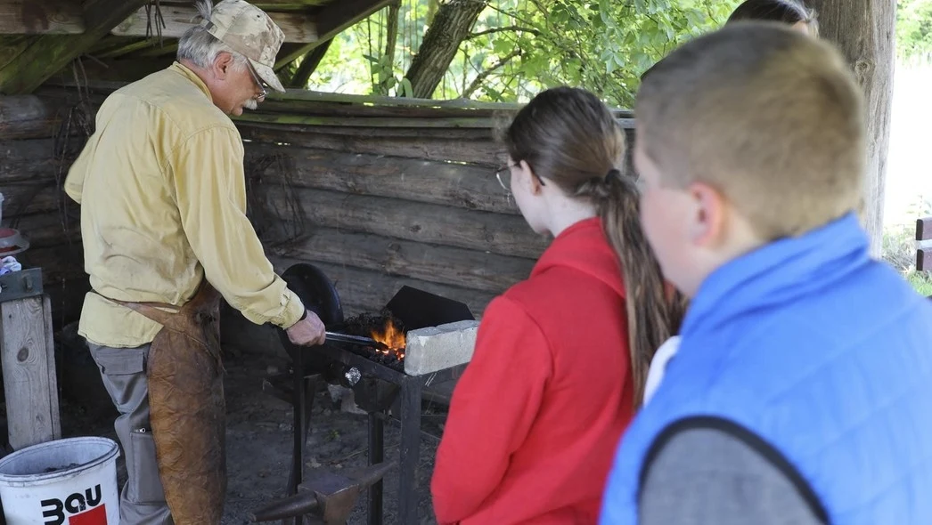 Starszy mężczyzna w nakryciu głowy i fartuchu skórzanym pracuje przy kowadle w kuźni na świeżym powietrzu, obserwowany przez grupę dzieci stojących bokiem; w tle drewniana szopa i płonący ogień w palenisku.