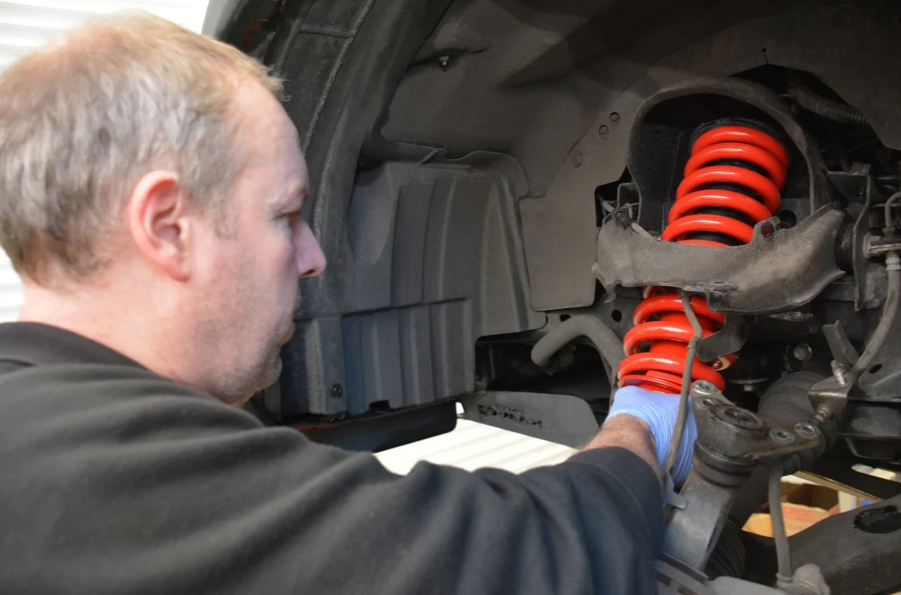 Objawy zużytych amortyzatorów - kiedy wymienić i ile to kosztuje? Mechanik montuje czerwony amortyzator spiralny w zawieszeniu samochodu, widoczna ręka w rękawiczce oraz elementy podwozia pojazdu.