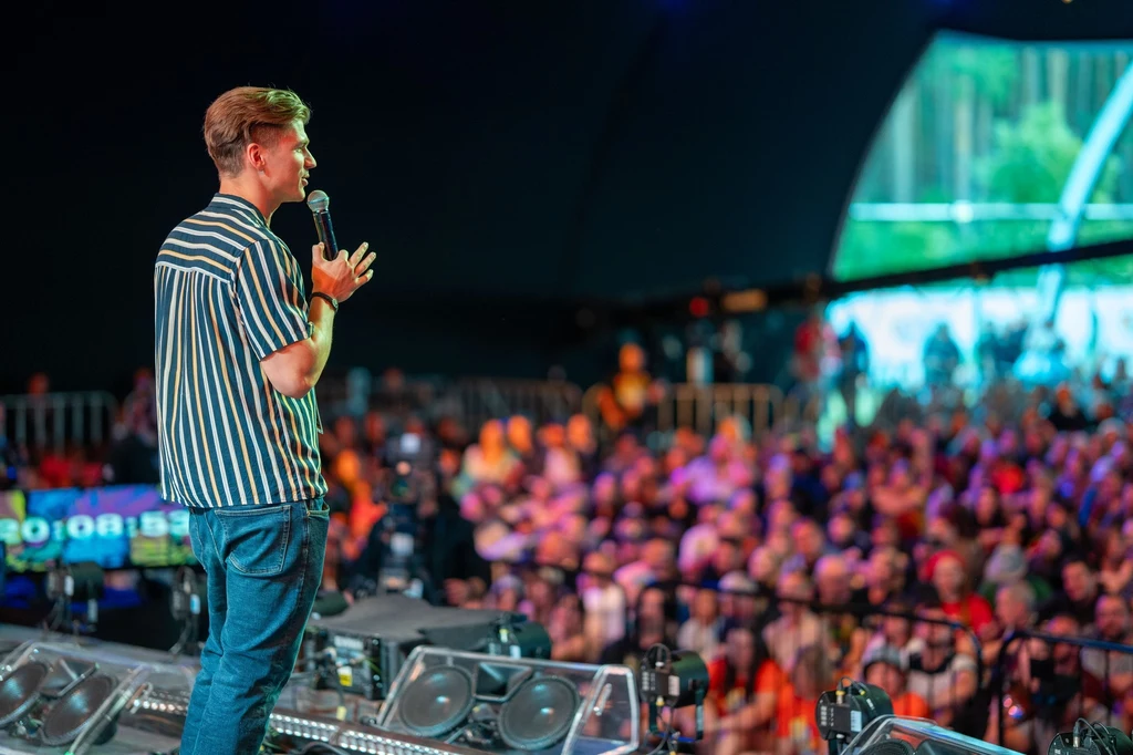 Stand-up "Nietykalni" (Kuba Rafalski i Roman Kostelecki) Mężczyzna w koszuli w paski trzyma mikrofon i przemawia do licznej, rozmytej publiczności zgromadzonej na wydarzeniu w dużej hali.
