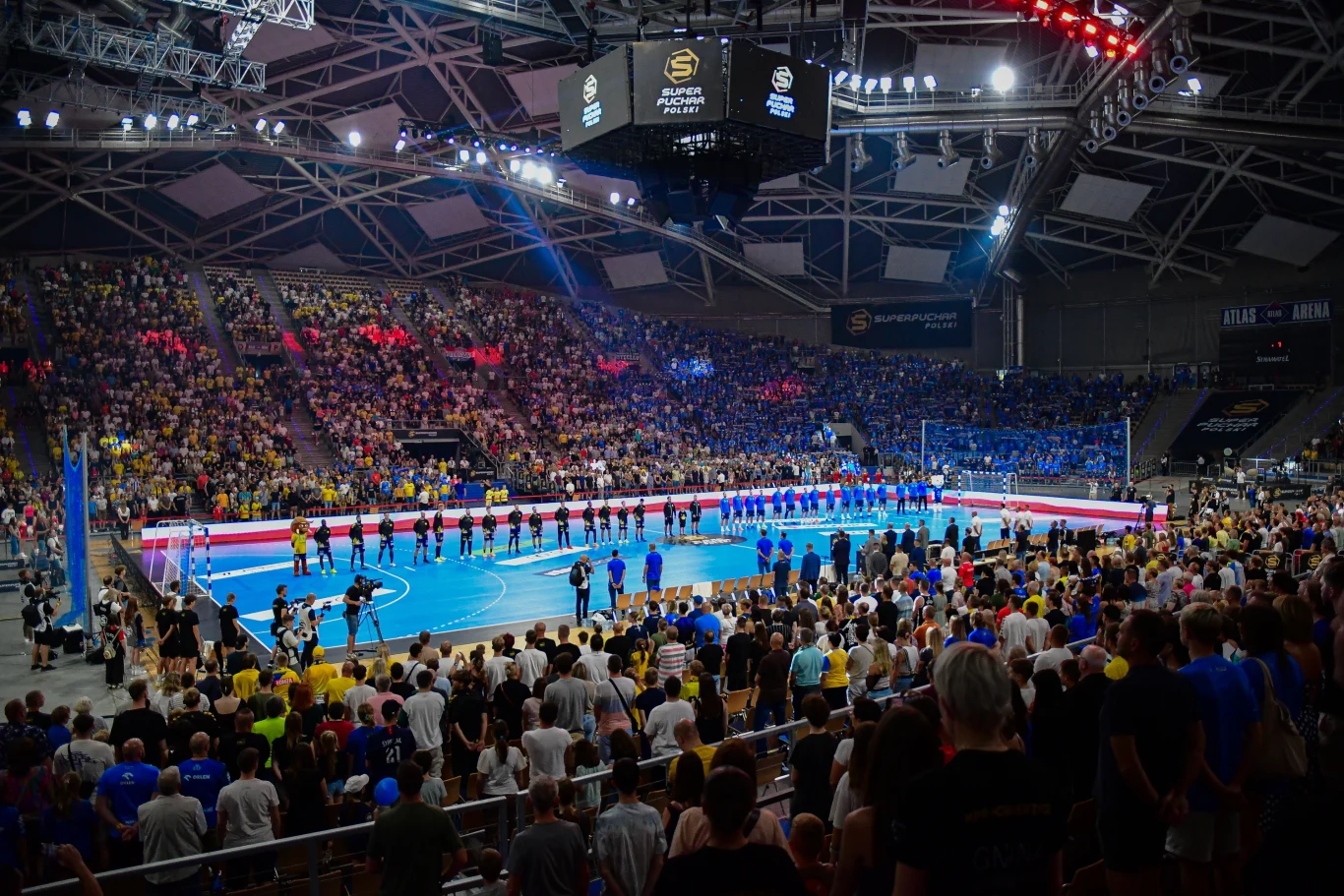 Tłum ludzi zgromadzony na dużej hali sportowej podczas meczu piłki ręcznej, zawodnicy ustawieni w rzędzie na środku boiska, intensywne światła i pełne trybuny podkreślają atmosferę ważnego wydarzenia sportowego.