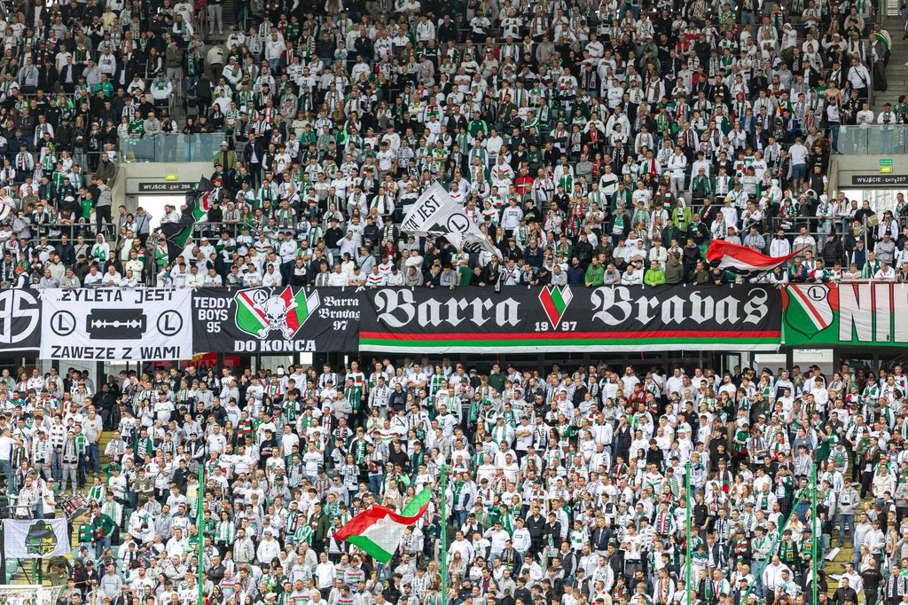 "Żyleta" - trybuna najzagorzalszych kibiców Legii Warszawa Tłum kibiców ubranych w barwy klubowe, uniesione flagi i transparenty, zapełnione trybuny stadionu podczas meczu piłkarskiego, widoczna energia i entuzjazm zgromadzonych fanów.
