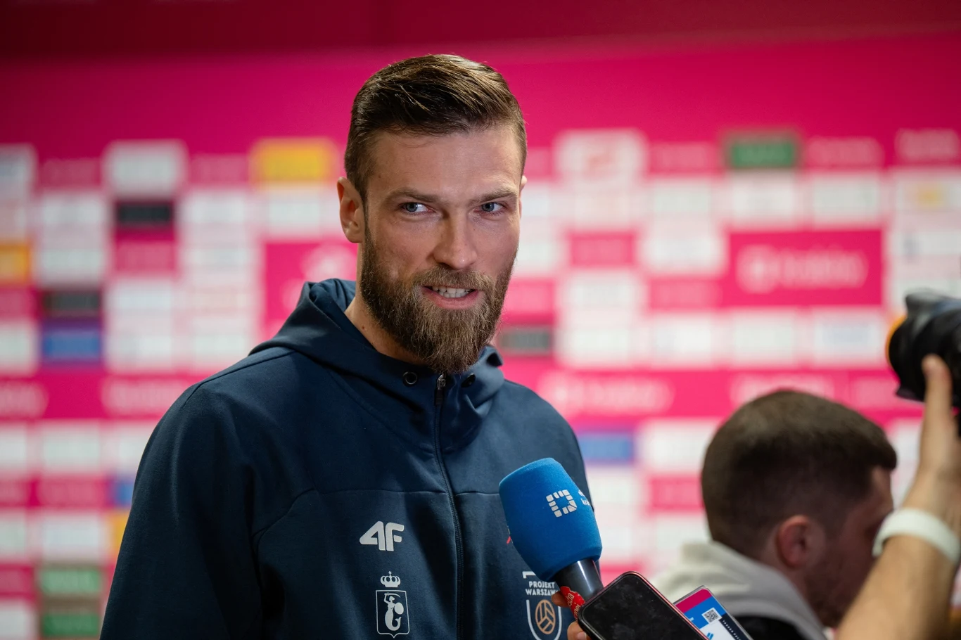 Mężczyzna z brodą w granatowej bluzie sportowej udziela wywiadu, stojąc przed ścianą z logotypami sponsorów, trzyma mikrofony różnych mediów.