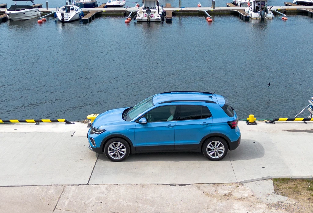Volkswagen T-Cross zaskakująco dobrze radzi sobie w długich trasach. To m.in. efekt całkiem przyzwoitego wygłuszenia kabiny Niebieski samochód typu SUV zaparkowany przy nabrzeżu w pobliżu portu z widocznymi łodziami i molo w tle.