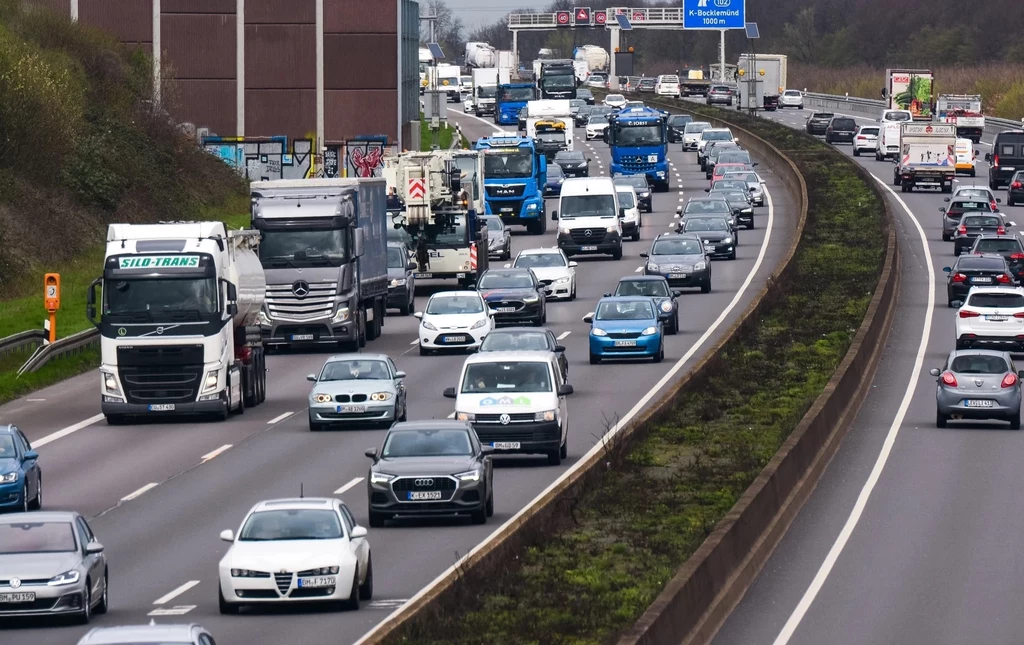Gęsty ruch samochodowy i ciężarówek na autostradzie, sznur pojazdów jadących w obu kierunkach z widocznymi znakami drogowymi i pasem zieleni rozdzielającym jezdnie.