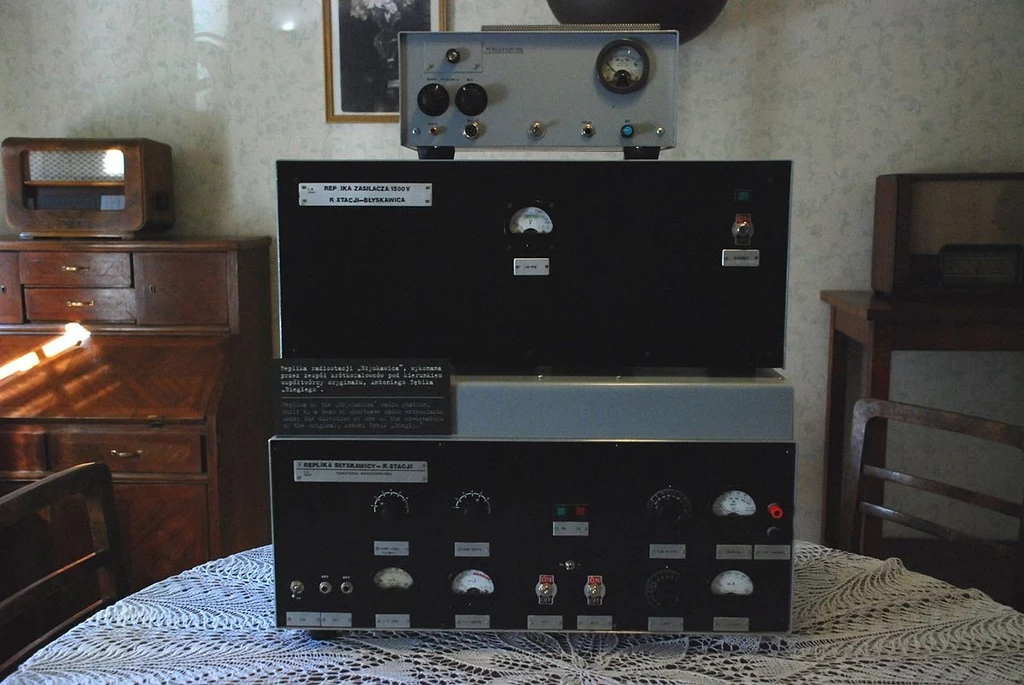 Duży aparat elektroniczny z licznymi wskaźnikami, pokrętłami i przyciskami ustawiony na stole przykrytym koronkowym obrusem, w tle drewniane meble i radioodbiornik.
