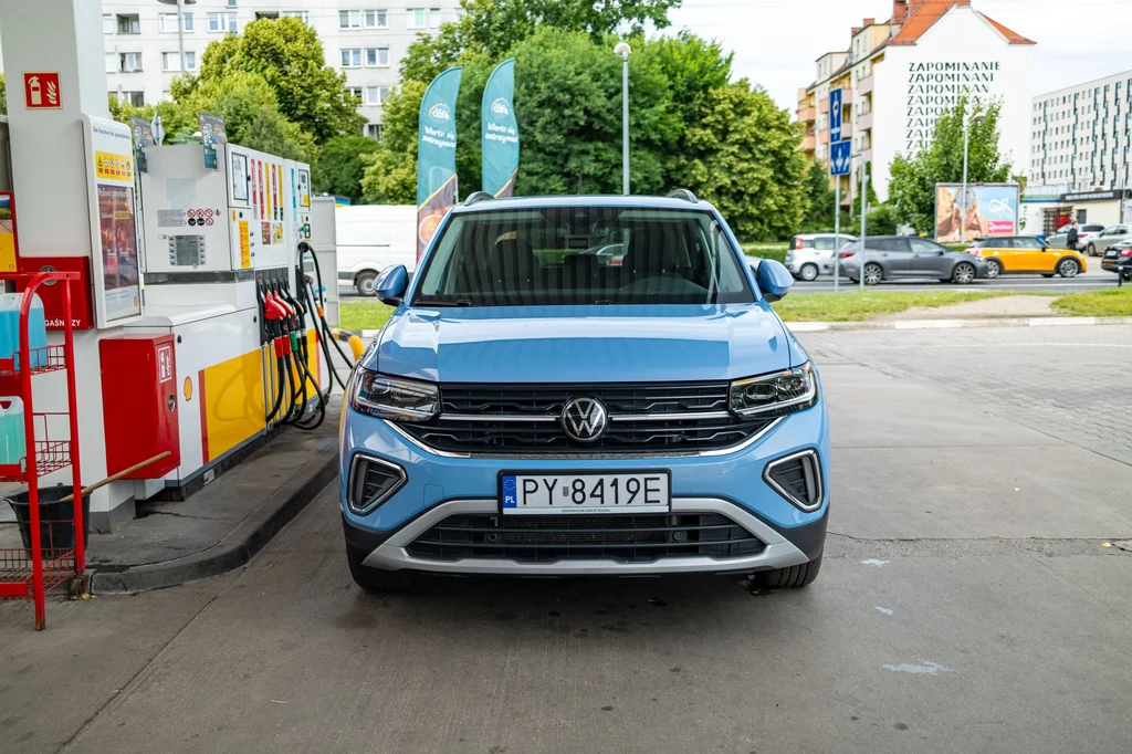 Średnie zużycie paliwa w naszym teście wahało się między 5 a 7 l/100 km Niebieski samochód marki Volkswagen stoi zaparkowany przy dystrybutorze paliw na stacji benzynowej, w tle widoczne budynki mieszkalne, zieleń oraz elementy stacji jak dystrybutory i tablice cenowe.