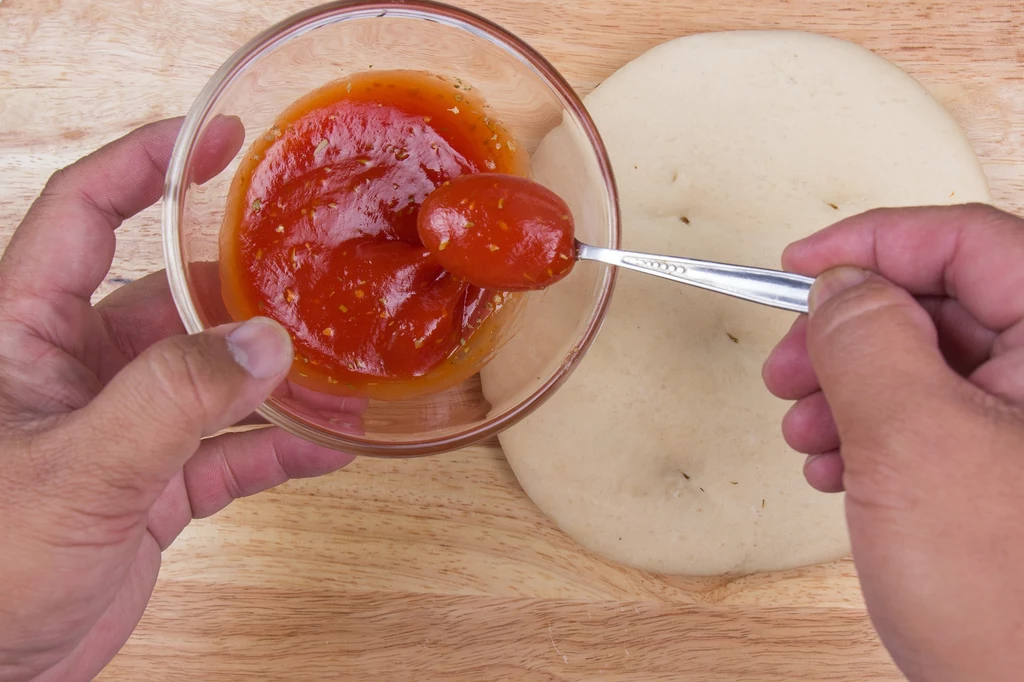 Ile likopenu zawierają pomidory, a ile ketchup? Odpowiedź zaskakuje Ręce osoby nakładającej sos pomidorowy z miseczki na surowe ciasto na pizze na drewnianym blacie.