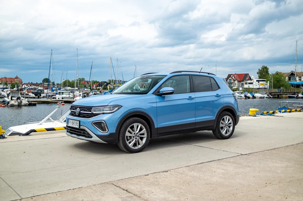 Volkswagenem T-Cross na drugi koniec Polski? Dlaczego nie? Ma duży bagażnik i jest całkiem nieźle wygłuszony Niebieski samochód osobowy Volkswagen T-Cross zaparkowany na nabrzeżu portowym z widokiem na cumujące jachty i łodzie oraz zabudowania w tle.