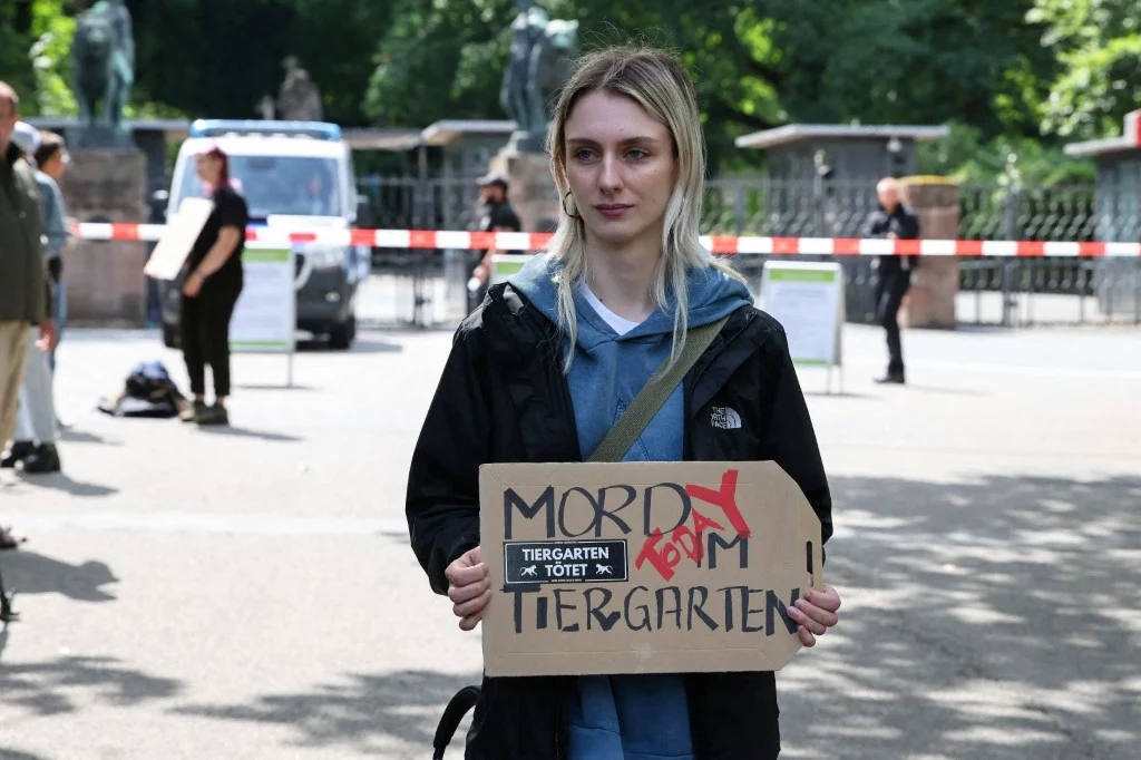 Młoda kobieta stoi na ulicy trzymając tekturowy transparent z napisem "Mord form Tiergarten", w tle widoczne są policyjne barierki oraz kilka osób, atmosfera sugeruje pokojowy protest w przestrzeni publicznej.