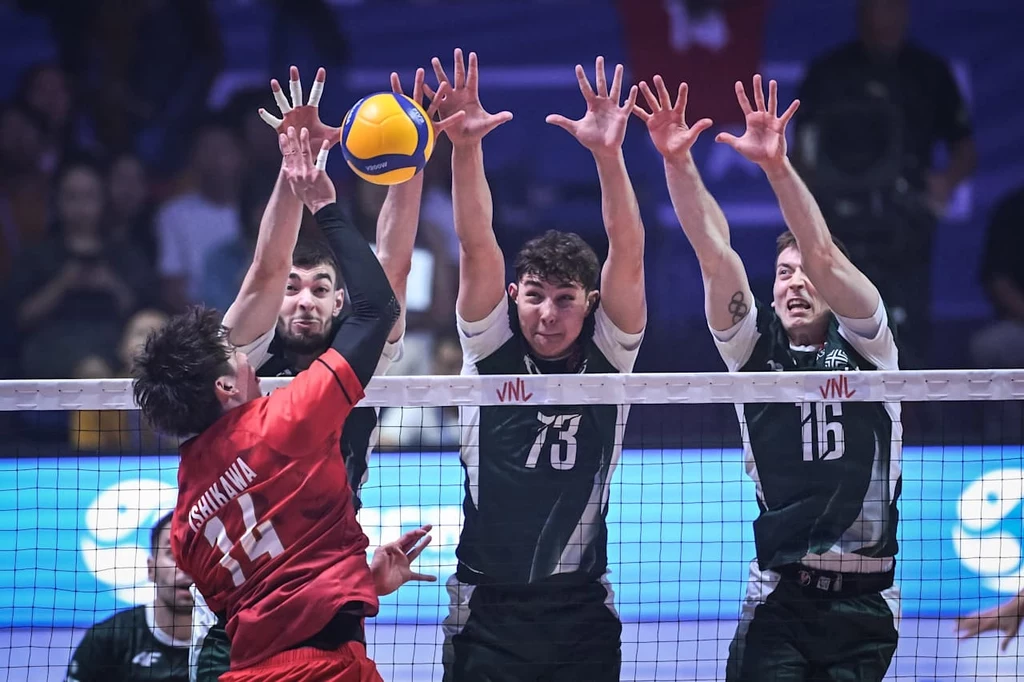 Drużyna w czarno-białych strojach próbuje zablokować atak zawodnika w czerwonym stroju przy siatce podczas meczu siatkówki, widoczna piłka oraz napięcie sportowe pomiędzy zawodnikami.