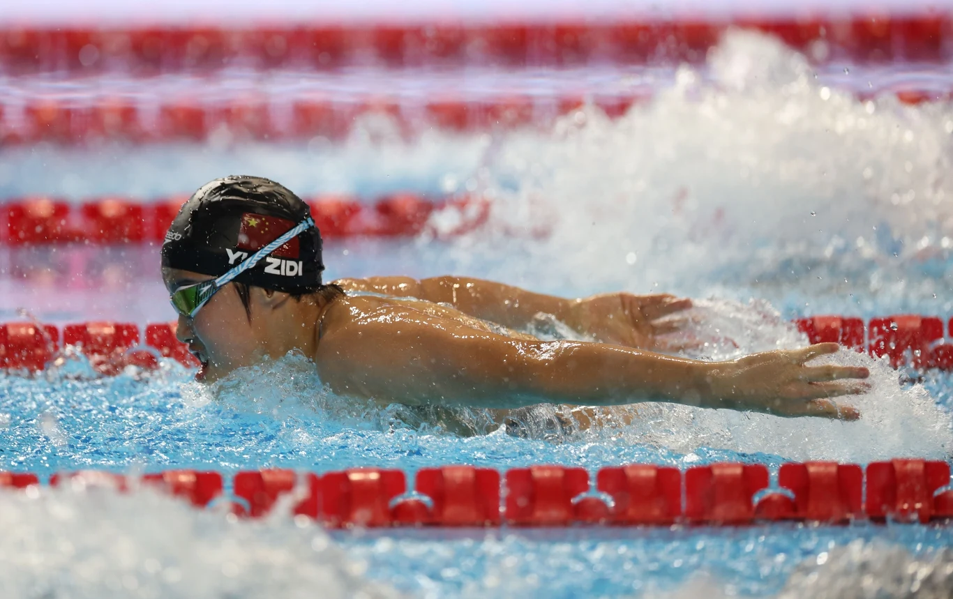 Pływak wykonujący styl motylkowy w basenie, otoczony falującą wodą i torami oddzielonymi czerwonymi płotkami; sportowiec nosi czepek i okularki, intensywnie skupiony na wyścigu