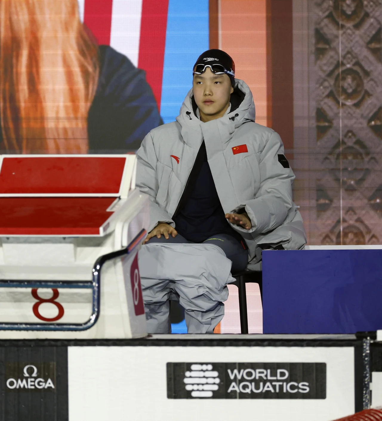 Zawodnik w sportowym stroju i okularach pływackich siedzi przy słupku startowym oznaczonym numerem 8, nosi jasną kurtkę z flagą Chin, w tle logo World Aquatics oraz kolorowa ściana.