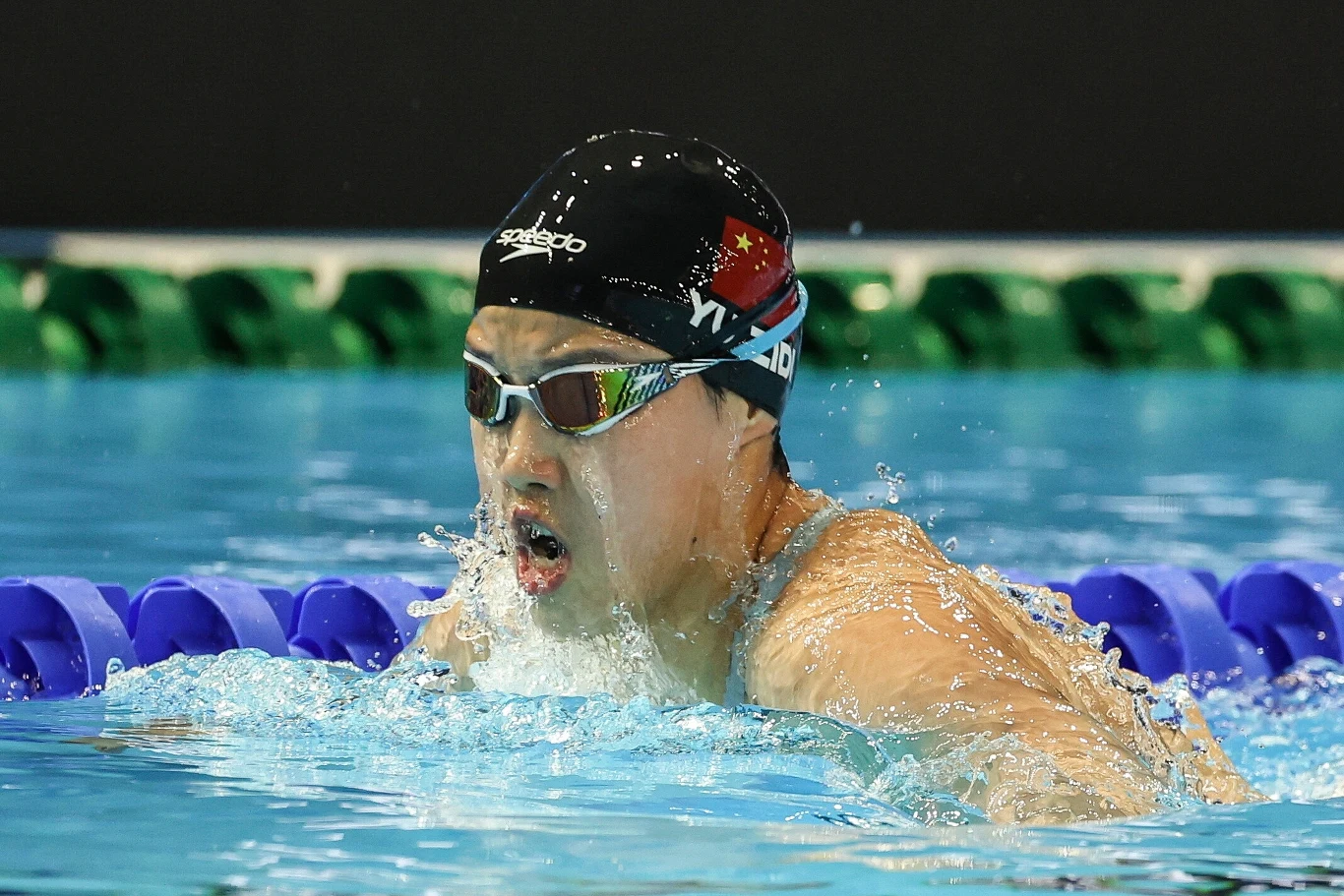 Sportowiec w czepku z flagą Chin i okularach pływackich wykonuje styl klasyczny podczas wyścigu na krytym basenie, wokół widoczne tory oddzielone niebieskimi bojkami.