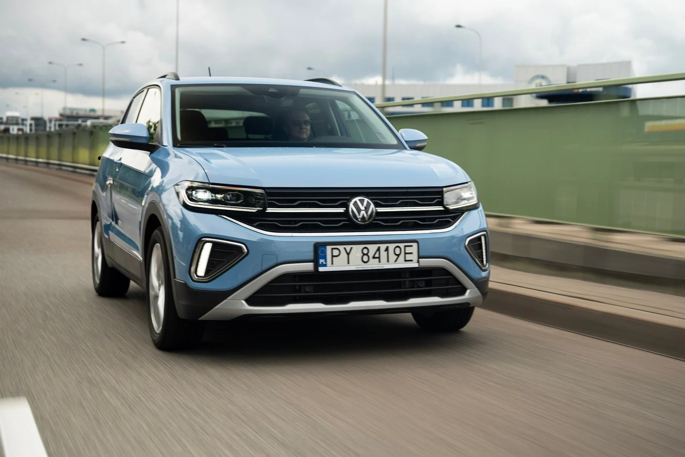 Volkswagen T-Cross przeszedł właśnie kurację odmładzającą Błękitny samochód marki Volkswagen jedzie po asfaltowej drodze, na tle zielonej barierki i nowoczesnych latarni, droga lekko rozmyta, podkreślająca ruch pojazdu.