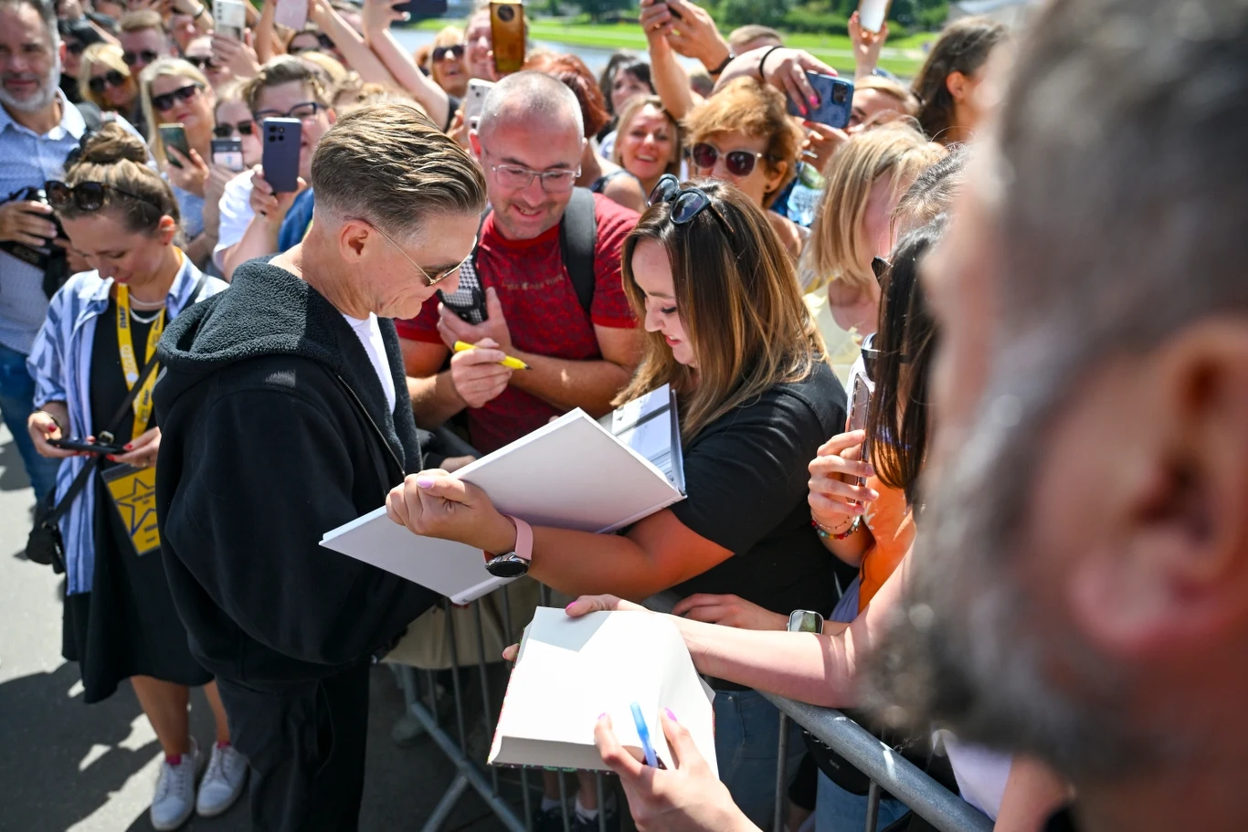 Bryan Adams pod Wawelem w Krakowie rozdawał autografy Tłum ludzi zgromadzonych przy barierce, wśród których znana osoba rozdaje autografy fanom, wokół wiele osób robi zdjęcia telefonami i okazuje entuzjazm.