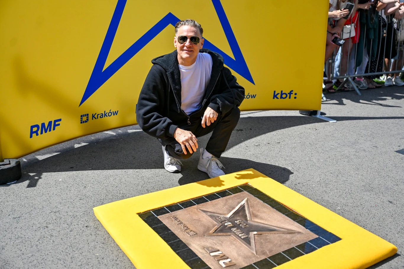 Bryan Adams ze swoją gwiazdą w Krakowie Mężczyzna w okularach przeciwsłonecznych i luźnej odzieży kuca przy tablicy z gwiazdą wmurowaną w chodnik, za nim znajdują się żółte tło promocyjne oraz widzowie za barierkami.