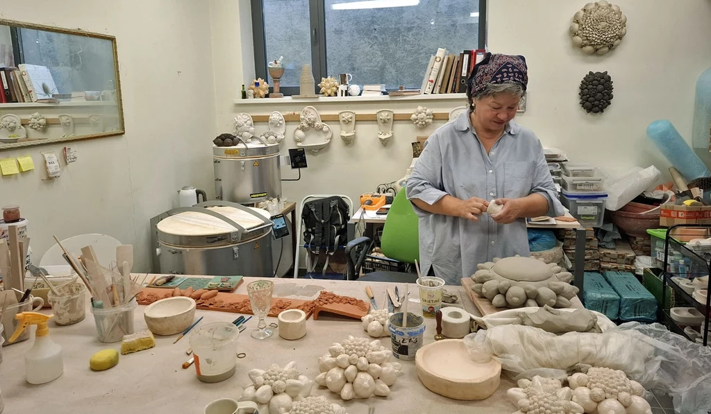 Artystka pracuje w jasnej pracowni ceramicznej, otoczona licznymi glinianymi rzeźbami oraz narzędziami do pracy z ceramiką; na stole znajdują się różnorodne formy i materiały związane z procesem tworzenia.
