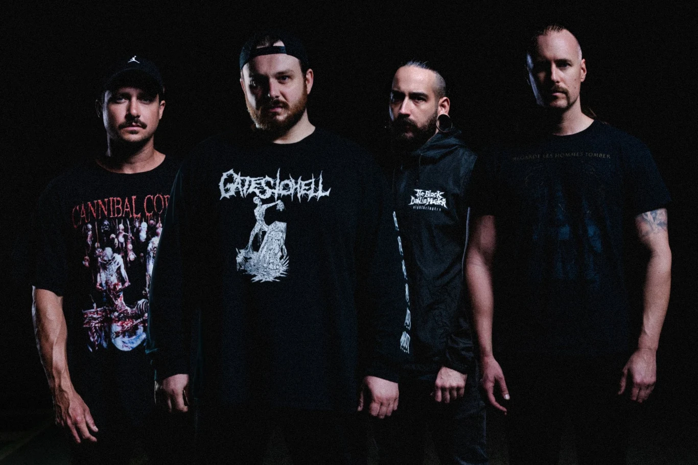 Aversions Crown w nowym składzie Czterech mężczyzn o poważnych wyrazach twarzy stoi obok siebie w ciemnym otoczeniu, ubranych w czarne bluzy i koszulki z nadrukami zespołów metalowych, światło podkreśla ich twarze i sylwetki.