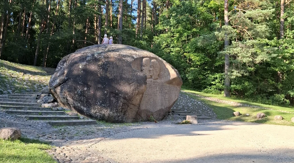 Ogromny głaz z wyrytym wizerunkiem i napisem znajduje się pośród zacienionych drzew, obok kamiennych schodów; dwoje ludzi stoi na szczycie głazu, reszta otoczenia tworzą leśne ścieżki i polana.