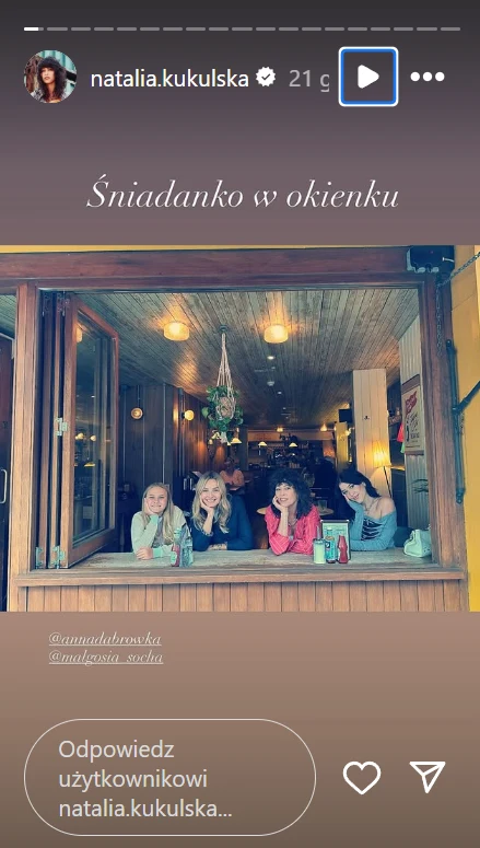 Cztery kobiety siedzą przy otwartym oknie kawiarni, uśmiechają się do obiektywu, wewnątrz widoczne drewniane wykończenia i ciepłe oświetlenie, tworzące przytulny klimat.