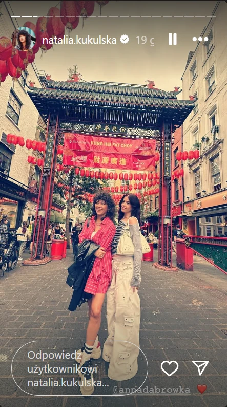 Dwie kobiety pozują razem na tle ozdobnej bramy w stylu azjatyckim udekorowanej czerwonymi lampionami i transparentem z napisem, wokół znajdują się miejskie budynki i przechodnie.