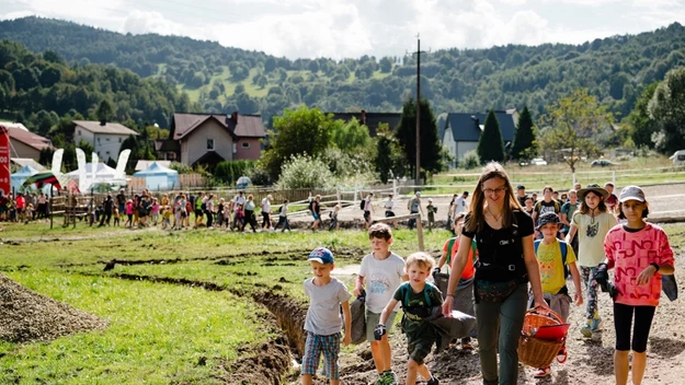Grupa dzieci wraz z opiekunką idzie ścieżką wśród zielonych terenów wiejskich, w oddali widać więcej osób, domy i zalesione wzgórza.
