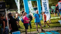 Grupa osób na imprezie plenerowej, na pierwszym planie kobieta z kijkami trekkingowymi i mężczyzna rozmawiają w pobliżu plansz informacyjnych i namiotu, w tle duże niebieskie dmuchane bramy oraz flagi promocyjne.