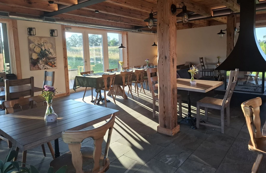 Jasno oświetlona sala restauracyjna z drewnianym sufitem i słupami, prosto ustawionymi stołami oraz krzesłami, ozdobiona bukietami kwiatów w wazonach, przy panoramicznych oknach wpuszczających światło słoneczne.