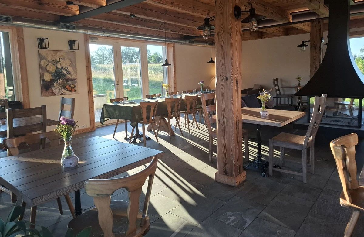 Przytulny salon z kominkiem i obszerny taras zachęcają do wspólnego spędzania czasu przy posiłkach Jasno oświetlona sala restauracyjna z drewnianym sufitem i słupami, prosto ustawionymi stołami oraz krzesłami, ozdobiona bukietami kwiatów w wazonach, przy panoramicznych oknach wpuszczających światło słoneczne.