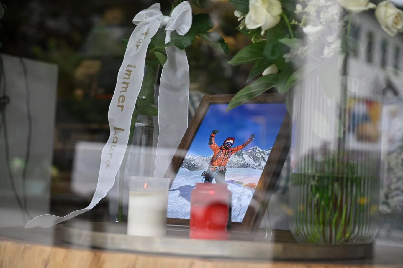 Fotografia ustawiona w ramce pokazuje osobę ubraną w kurtkę zimową na tle ośnieżonych gór, przed ramką ustawione są zapalone świece oraz bukiet białych kwiatów z białą wstążką z napisem.