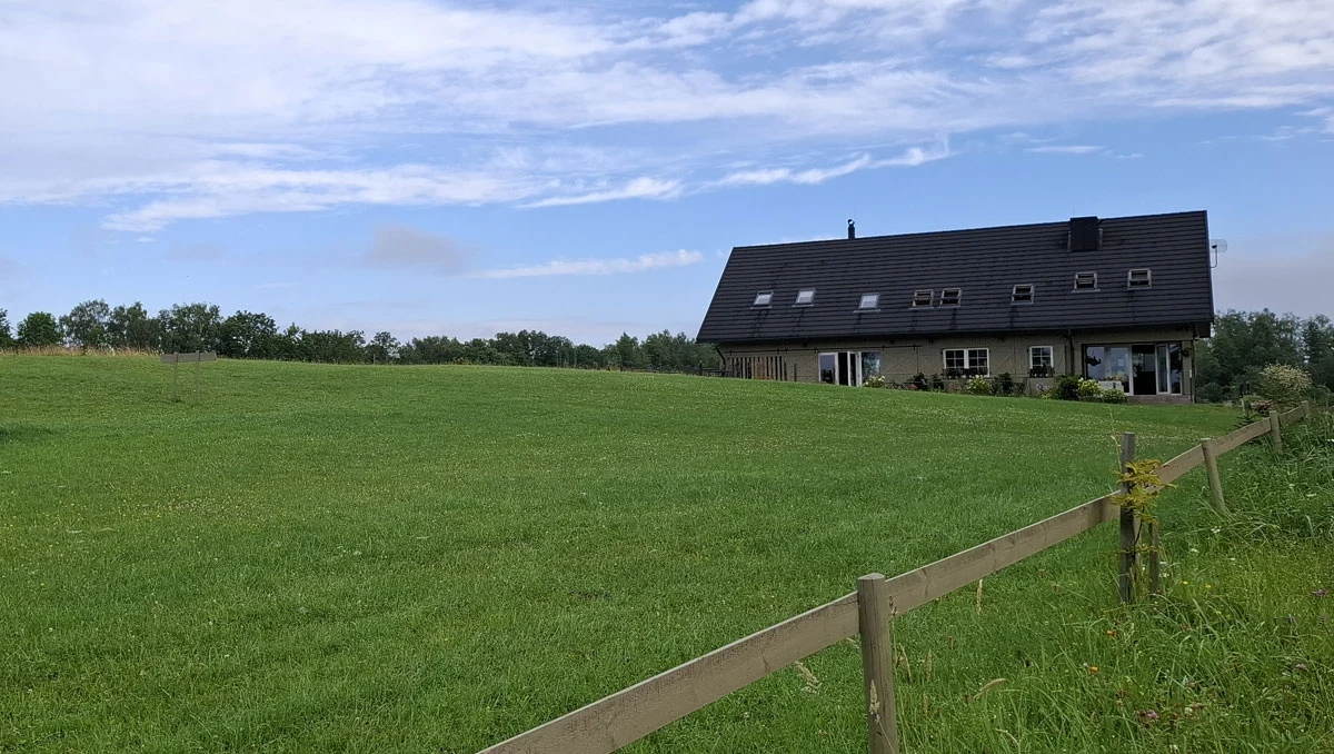 Szwajcarska para zafascynowana litewską przyrodą prrzerobiła opuszczoną stodołę na komfortowy dom goscinny Nowoczesny budynek mieszkalny z dużym, zadbanym trawnikiem otoczonym drewnianym ogrodzeniem, na tle nieba z lekkim zachmurzeniem i linią drzew w oddali.