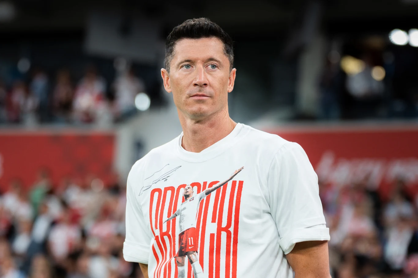 Robert Lewandowski Mężczyzna o krótkich ciemnych włosach ubrany w białą koszulkę z nadrukiem, stojący na tle rozmytej publiczności na stadionie sportowym.