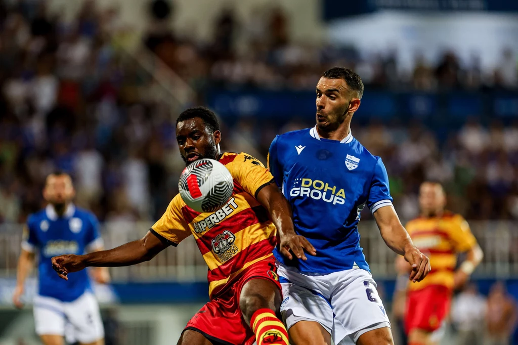 Mecz Jagiellonia - Novi Pazar Dwóch piłkarzy rywalizuje o piłkę podczas meczu, jeden w żółto-czerwonym stroju, drugi w niebiesko-białym, akcja toczy się na tle rozmytego tłumu kibiców.