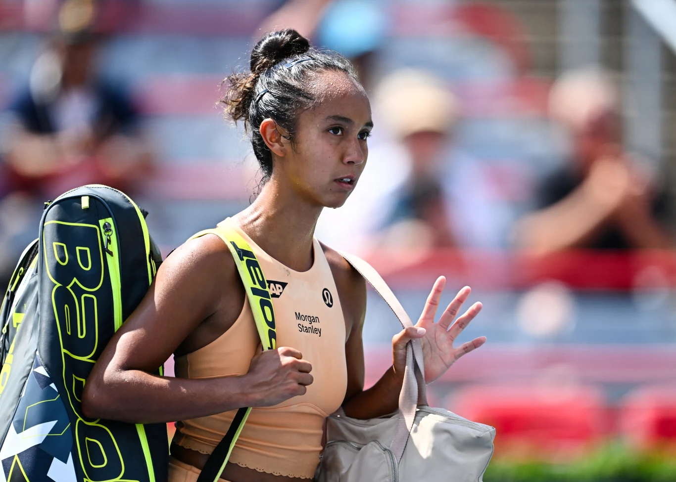 Tenisistka z torbą sportową na ramieniu przechodzi obok trybun, ujęcie wykonane podczas rozgrywek na kortach, widoczne logo firmy sportowej na rakiecie, sportowa atmosfera oraz skupienie na twarzy zawodniczki.