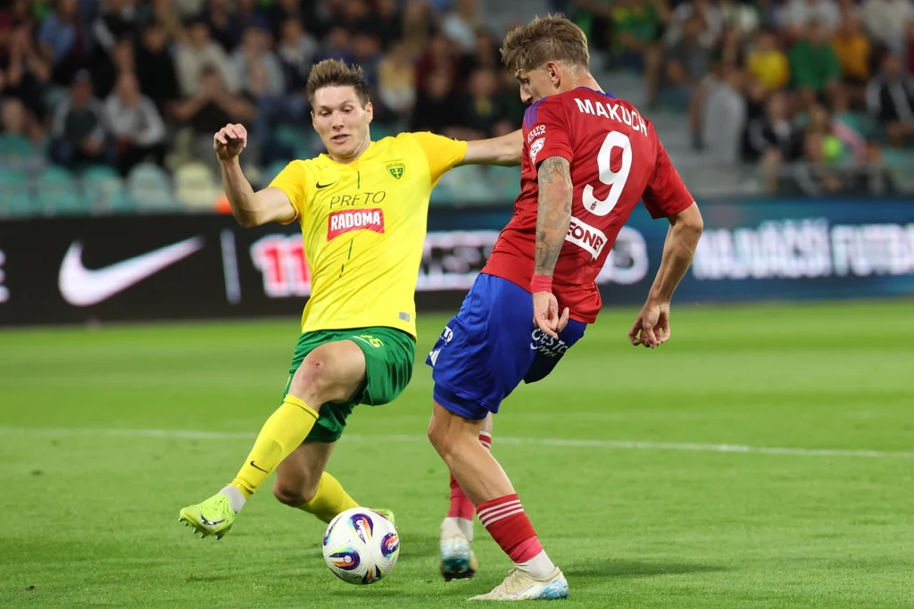 Dwóch piłkarzy w trakcie dynamicznej walki o piłkę na murawie stadionu, zawodnik w żółtej koszulce wykonuje wślizg próbując odebrać piłkę rywalowi w czerwonym stroju, w tle rozmazana publiczność na trybunach.