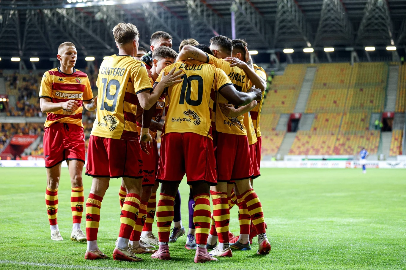 Grupa piłkarzy w żółto-czerwonych strojach obejmuje się na murawie stadionu po zdobytej bramce, wokół widać puste trybuny i światła stadionu.