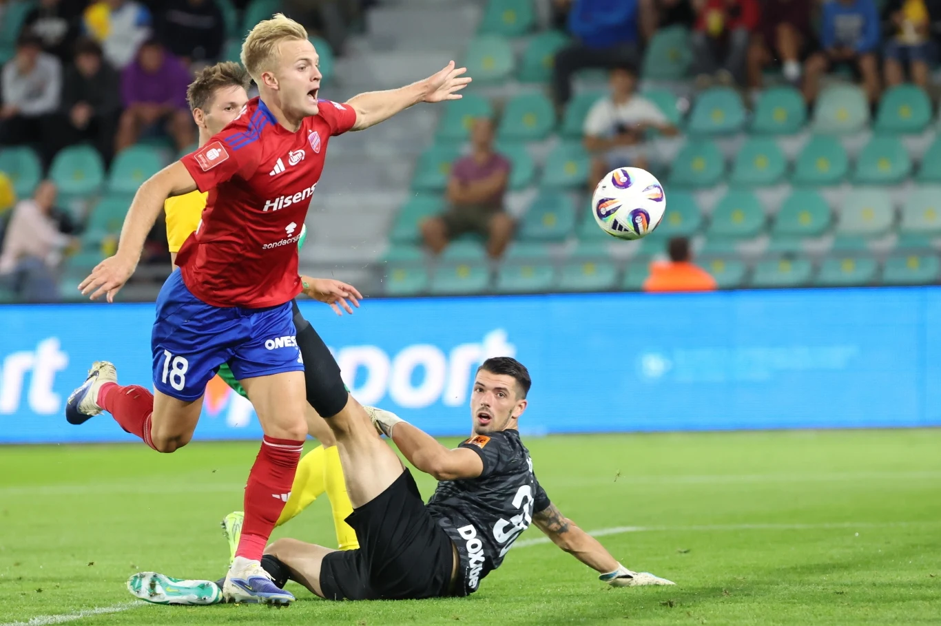 Raków awansował do 3. rundy eliminacji Ligi Konferencji Piłkarz w czerwono-niebieskim stroju przewraca się podczas walki o piłkę tuż przed bramkarzem w czarnym stroju, wokół nich znajduje się inny zawodnik oraz trybuny pełne publiczności.