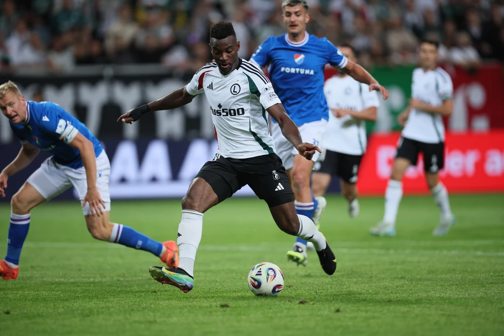 Legia Warszawa - Banik Ostrawa Piłkarz w biało-czarnym stroju drybluje piłkę podczas meczu, ścigany przez przeciwników w niebieskich oraz białych koszulkach, na tle rozmytej publiczności na stadionie.