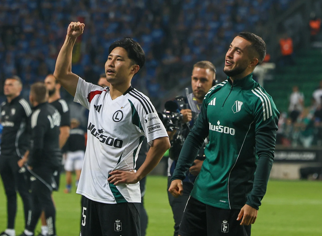 Ryoya Morishita i Wahan Biczachczjan, 31.07.2025 r. Dwóch piłkarzy w strojach klubu Legia Warszawa stoi na murawie stadionu, jeden unosi rękę w geście triumfu, drugi uśmiecha się, w tle znajduje się reszta drużyny i rozmyty tłum kibiców na trybunach.